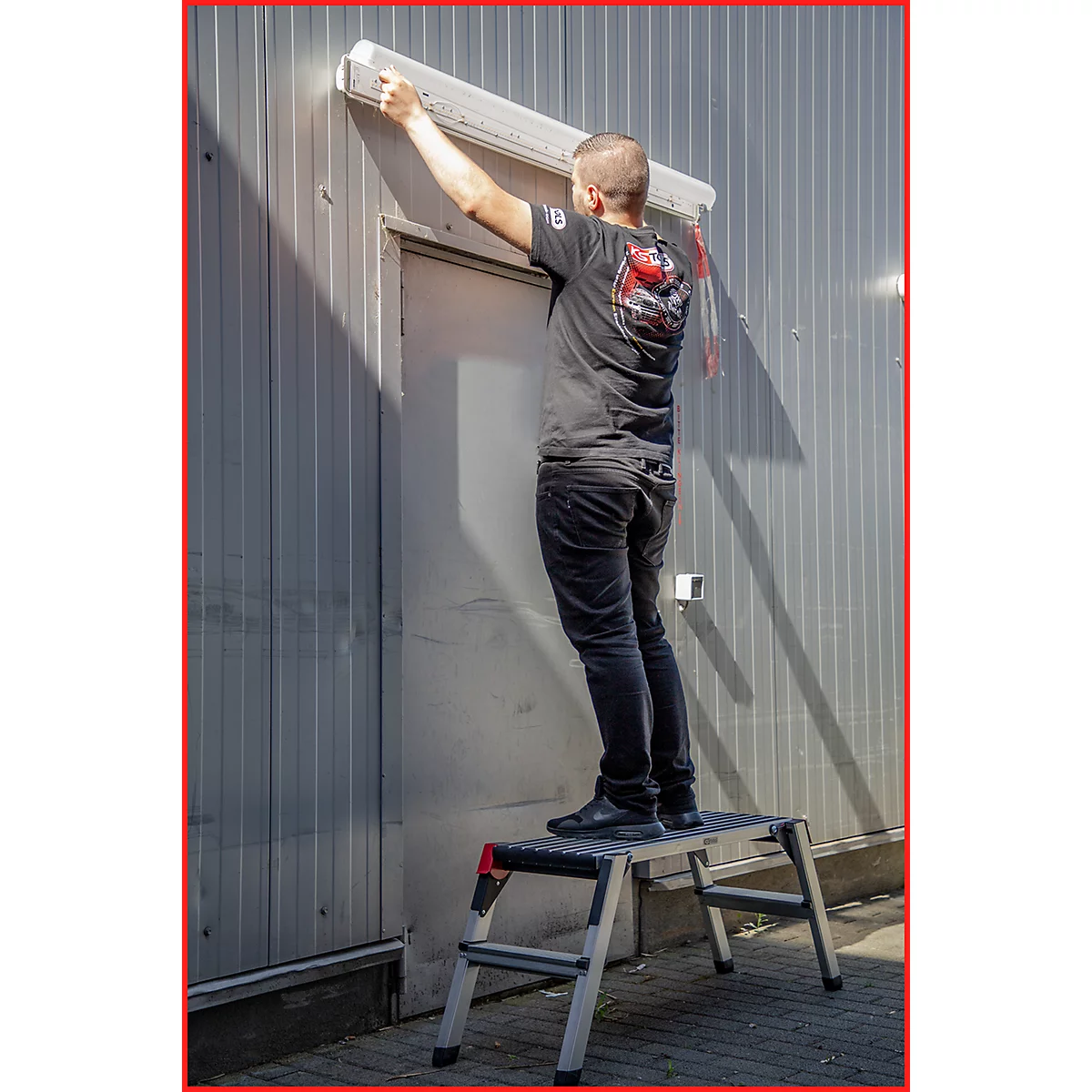 Ein Mann installiert eine Markise über einer Tür; er steht auf einer Trittleiter. Die Tür ist in eine graue Metallwand eingelassen.